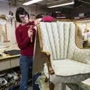 A young female upholsterer using a tack hammer on a chair in an upholstery shop.