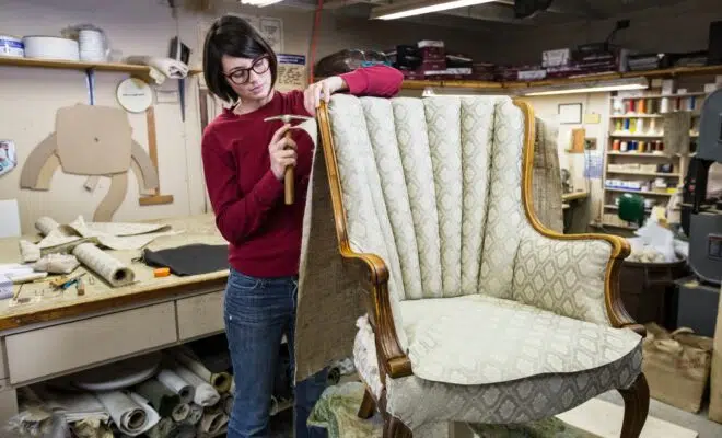 A young female upholsterer using a tack hammer on a chair in an upholstery shop.