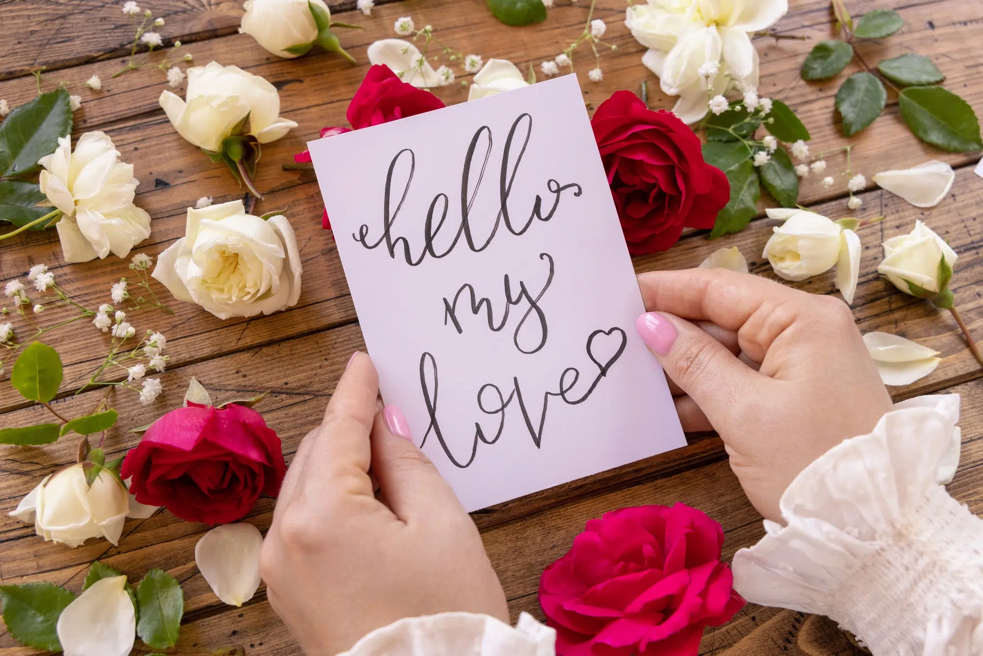 Hands with card HELLO MY LOVE near red and cream flowers close up on a wooden table